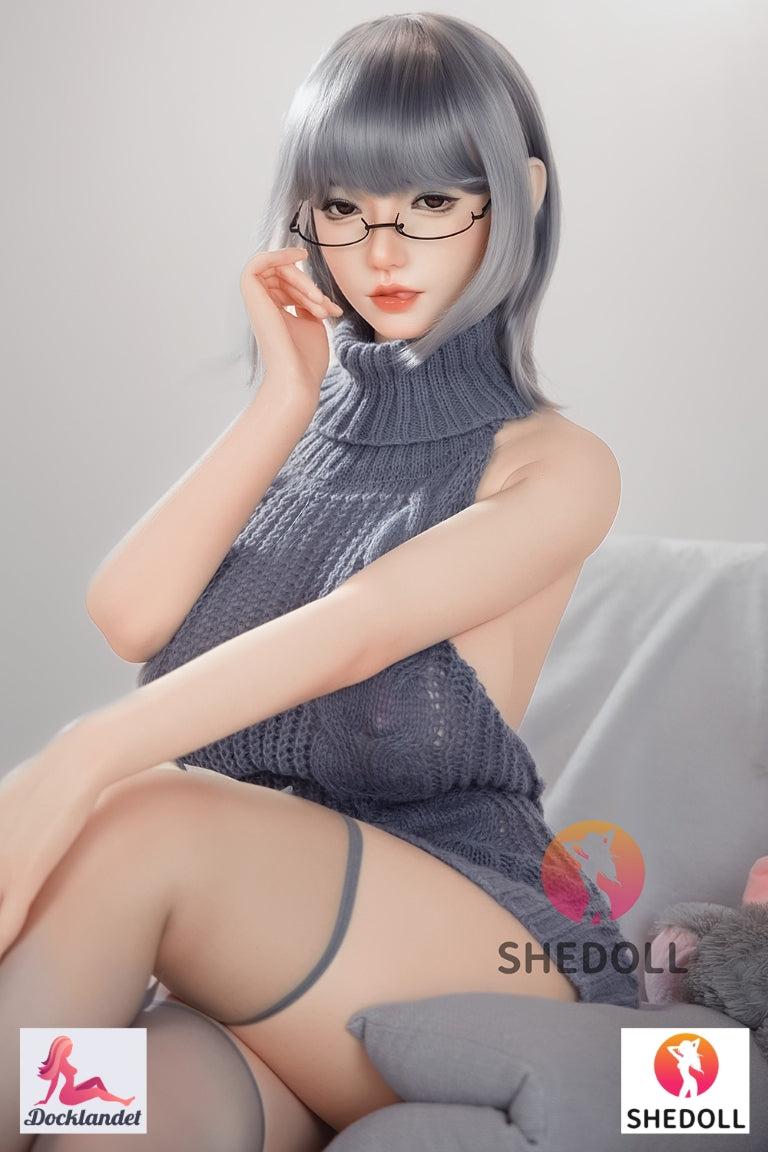 Shedoll sexdocka med kort grå frisyr och glasögon i grå stickad klänning sittandes i soffa bredvid kanin-gosedjur – realistisk silikon-docka från Docklandet