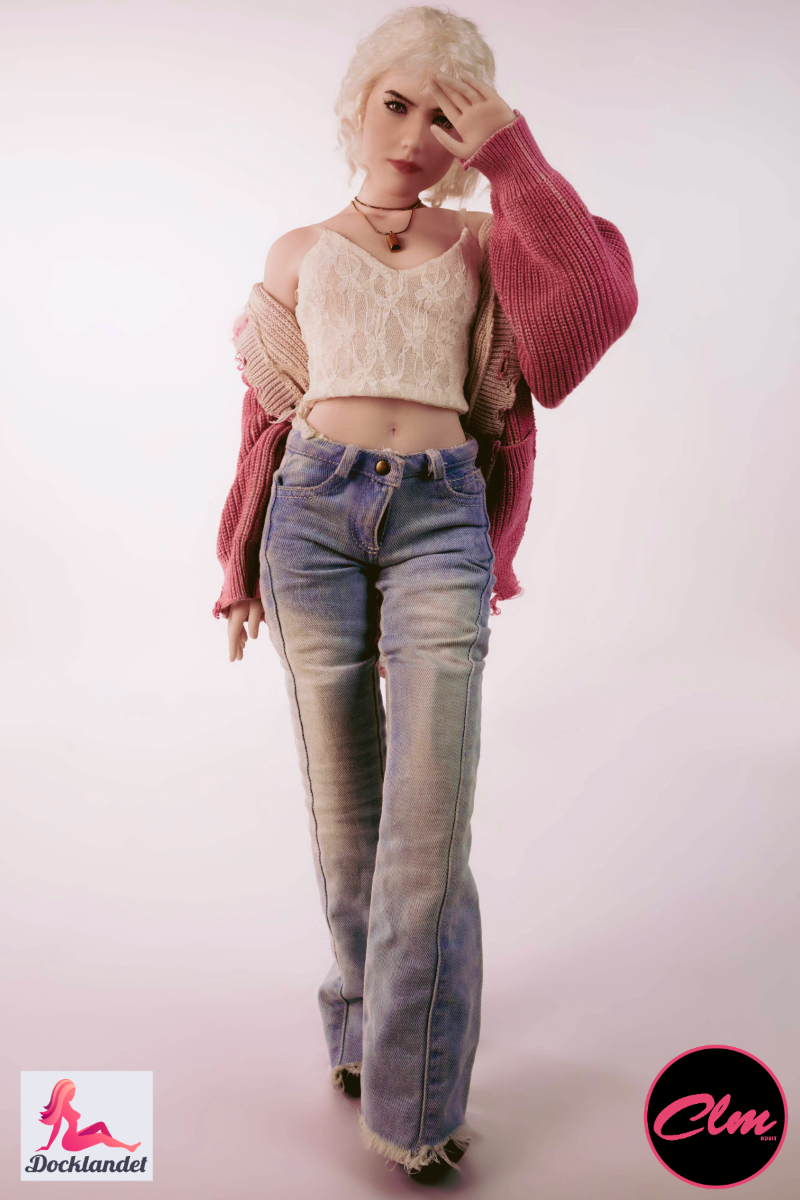 Sexdocka Tara realistisk mini 60 cm i silikon, blond docka i jeans, kort topp och rosa jacka, står upprätt mot ljus studiobakgrund.